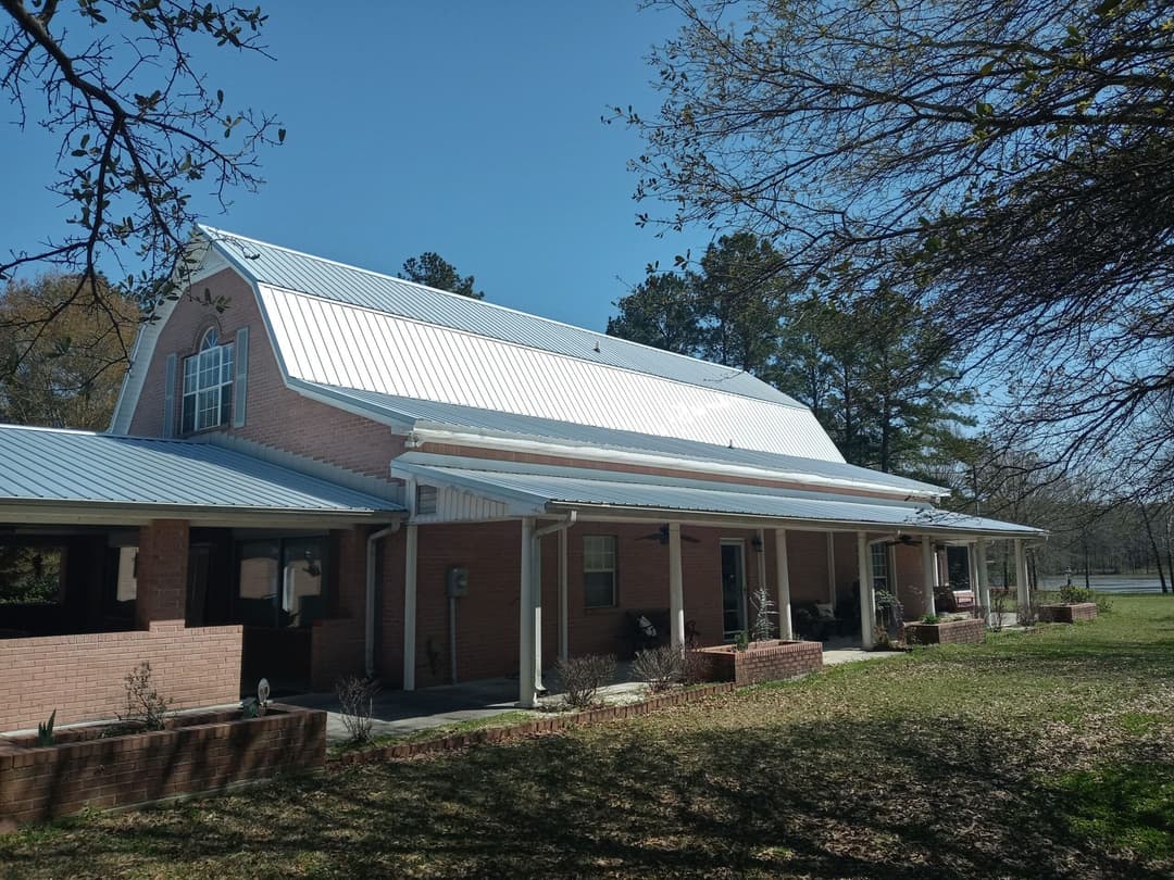 Charming brick house with a metal roof, surrounded by trees and greenery on a sunny day.